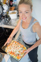 Image showing Smiling young healthy woman holding and proudly showing glass baking try with row vegetarian dish ingredients before putting it into oven.