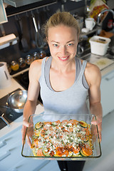 Image showing Smiling young healthy woman holding and proudly showing glass baking try with row vegetarian dish ingredients before putting it into oven.