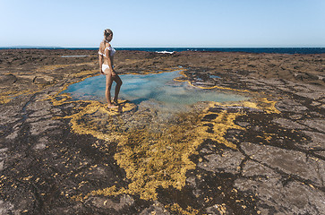 Image showing Woman stands by a small rock pool edged with yellow seaweed