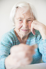 Image showing Sad elderly woman sitting at the table at home and looking miserably at only remaining coin from pension in her hand.