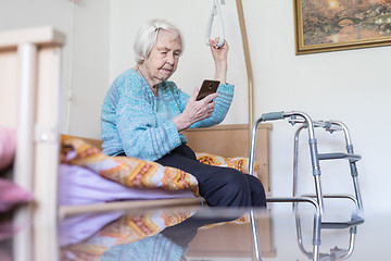 Image showing Elderly 96 years old woman reading phone message while sitting on medical bed supporting her by holder.