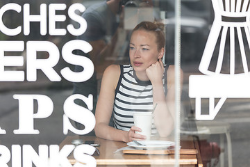 Image showing Young caucasian woman sitting alone in coffee shop thoughtfully leaning on her hand, looking trough the window