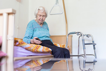 Image showing Elderly 96 years old woman reading phone message while sitting on medical bed supporting her by holder.