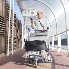 Image showing Young casual woman using mobile phone application while transporting luggage from arrival parking to international airport departure termainal by luggage trolley