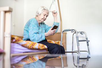 Image showing Elderly 96 years old woman reading phone message while sitting on medical bed supporting her by holder.