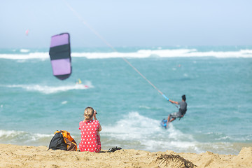 Image showing Girl sitting on sandy beach watching kite surfer in the sea.