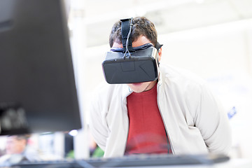 Image showing Young man wearing virtual reality headset and gesturing while sitting at his desk in creative office