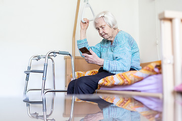 Image showing Elderly 96 years old woman reading phone message while sitting on medical bed supporting her by holder.