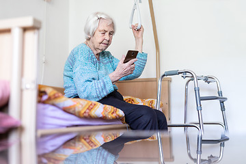 Image showing Elderly 96 years old woman reading phone message while sitting on medical bed supporting her by holder.