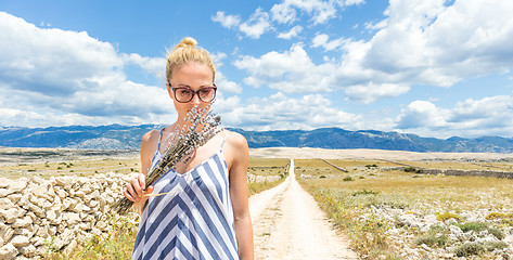 Image showing Woman in summer dress holding and smelling bouquet of lavender flowers while walking outdoor through dry rocky Mediterranean Croatian coast lanscape on Pag island in summertime