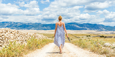 Image showing Rear view of woman in summer dress holding bouquet of lavender flowers while walking outdoor through dry rocky Mediterranean Croatian coast lanscape on Pag island in summertime