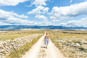 Image showing Caucasian young woman in summer dress holding bouquet of lavender flowers while walking outdoor through dry rocky Mediterranean Croatian coast lanscape on Pag island in summertime