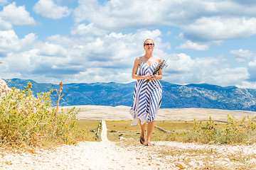 Image showing Caucasian young woman in summer dress holding bouquet of lavender flowers while walking outdoor through dry rocky Mediterranean Croatian coast lanscape on Pag island in summertime