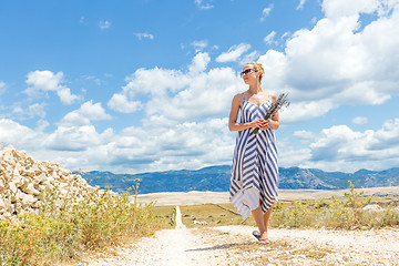 Image showing Caucasian young woman in summer dress holding bouquet of lavender flowers while walking outdoor through dry rocky Mediterranean Croatian coast lanscape on Pag island in summertime