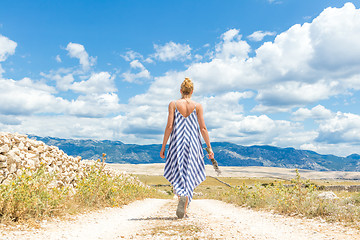 Image showing Rear view of woman in summer dress holding bouquet of lavender flowers while walking outdoor through dry rocky Mediterranean Croatian coast lanscape on Pag island in summertime