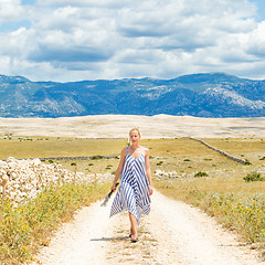 Image showing Caucasian young woman in summer dress holding bouquet of lavender flowers while walking outdoor through dry rocky Mediterranean Croatian coast lanscape on Pag island in summertime