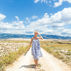 Image showing Caucasian young woman in summer dress holding bouquet of lavender flowers while walking outdoor through dry rocky Mediterranean Croatian coast lanscape on Pag island in summertime