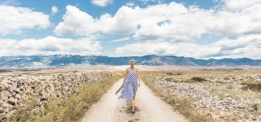 Image showing Caucasian young woman in summer dress holding bouquet of lavender flowers while walking outdoor through dry rocky Mediterranean Croatian coast lanscape on Pag island in summertime