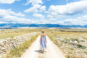 Image showing Caucasian young woman in summer dress holding bouquet of lavender flowers while walking outdoor through dry rocky Mediterranean Croatian coast lanscape on Pag island in summertime