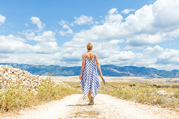 Image showing Rear view of woman in summer dress holding bouquet of lavender flowers while walking outdoor through dry rocky Mediterranean Croatian coast lanscape on Pag island in summertime