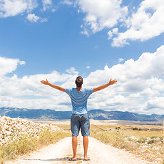 Image showing Rear view of casual sporty man standing on a dirt country road rising hands up to the clouds on a blue summer sky. Freedom and travel adventure concept.