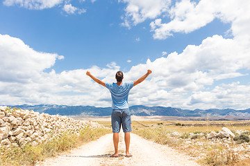 Image showing Rear view of casual sporty man standing on a dirt country road rising hands up to the clouds on a blue summer sky. Freedom and travel adventure concept.