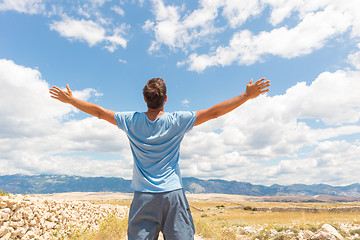 Image showing Rear view of casual sporty man standing on a dirt country road rising hands up to the clouds on a blue summer sky. Freedom and travel adventure concept.