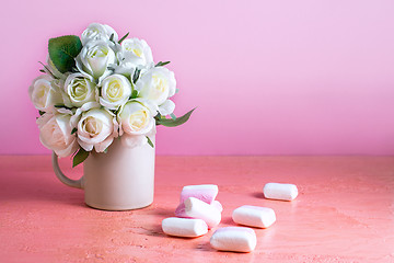 Image showing Small white and pink marshmallows are scattered on a pale pink background next to a vase of roses. Place for text.