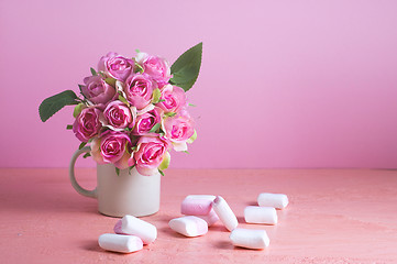 Image showing Small white and pink marshmallows are scattered on a pale pink background next to a vase of roses. Place for text.