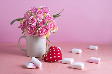 Image showing Small white and pink marshmallows are scattered on a pale pink background next to a vase of roses. Place for text.