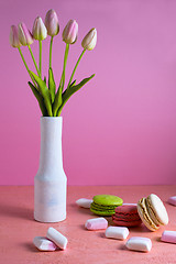 Image showing Macaroons and small white and pink marshmallows are scattered on a pale pink background next to a vase of tulips. Place for text.