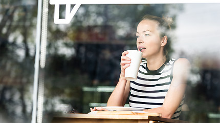 Image showing Young caucasian woman sitting alone in coffee shop thoughtfully leaning on her hand, looking trough the window