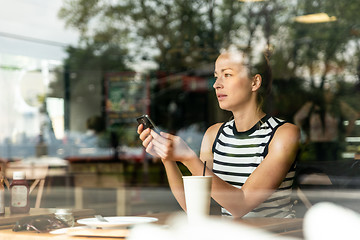 Image showing Thoughtful caucasian woman holding mobile phone while looking through the coffee shop window during coffee break.