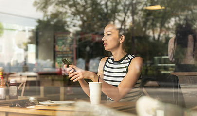 Image showing Thoughtful caucasian woman holding mobile phone while looking through the coffee shop window during coffee break.