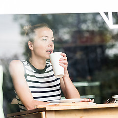 Image showing Young caucasian woman sitting alone in coffee shop thoughtfully leaning on her hand, looking trough the window