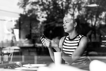 Image showing Thoughtful caucasian woman holding mobile phone while looking through the coffee shop window during coffee break.