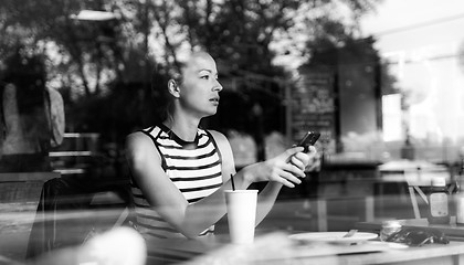 Image showing Thoughtful caucasian woman holding mobile phone while looking through the coffee shop window during coffee break.