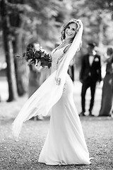 Image showing Full length portrait of beautiful sensual young blond bride in long white wedding dress and veil, holding bouquet outdoors in natural background.