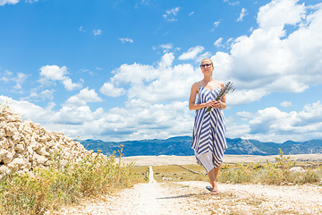 Image showing Caucasian young woman in summer dress holding bouquet of lavender flowers while walking outdoor through dry rocky Mediterranean Croatian coast lanscape on Pag island in summertime