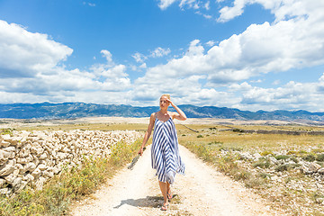 Image showing Caucasian young woman in summer dress holding bouquet of lavender flowers while walking outdoor through dry rocky Mediterranean Croatian coast lanscape on Pag island in summertime