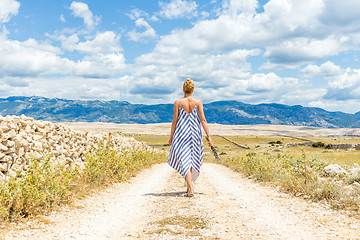 Image showing Rear view of woman in summer dress holding bouquet of lavender flowers while walking outdoor through dry rocky Mediterranean Croatian coast lanscape on Pag island in summertime