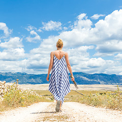Image showing Rear view of woman in summer dress holding bouquet of lavender flowers while walking outdoor through dry rocky Mediterranean Croatian coast lanscape on Pag island in summertime