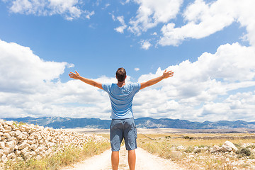 Image showing Rear view of casual sporty man standing on a dirt country road rising hands up to the clouds on a blue summer sky. Freedom and travel adventure concept.