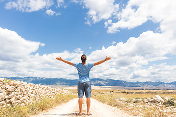 Image showing Rear view of casual sporty man standing on a dirt country road rising hands up to the clouds on a blue summer sky. Freedom and travel adventure concept.