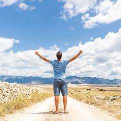 Image showing Rear view of casual sporty man standing on a dirt country road rising hands up to the clouds on a blue summer sky. Freedom and travel adventure concept.