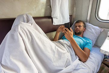 Image showing A man lies on the bottom shelf in a train car with a phone in his hands