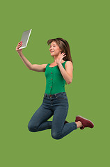 Image showing Image of young woman over green background using laptop computer or tablet gadget while jumping.