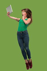 Image showing Image of young woman over green background using laptop computer or tablet gadget while jumping.