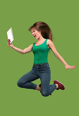 Image showing Image of young woman over green background using laptop computer or tablet gadget while jumping.