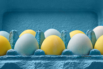 Image showing Close-up multicolored painted in yellow blue eggs in a blue cardboard box around blue background with copy space. Easter composition.